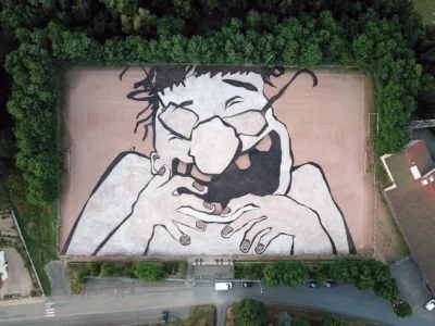 Aerial view of a massive black-and-white portrait painted across a sports field, depicting a laughing person with glasses and hands beneath their chin, surrounded by dense tree
