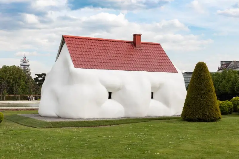 Erwin Wurm's 'Fat House' sculpture, a white cottage with swollen, bulging walls and a red tiled roof installed on a manicured lawn.
