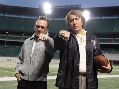 Left Christian Bale and right Nicholas Cage pose on a football field inside a large stadium, extending championship rings toward the camera. One wears sunglasses and a gray knit jacket, while the other wears a black Raiders jacket, holds a brown football, and displays a large ring under bright stadium light