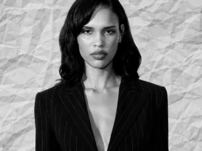 Editorial portrait of Nara Aziza Smith wearing a black pinstripe blazer with a deep neckline, standing against a textured gray backdrop and looking directly into the camera