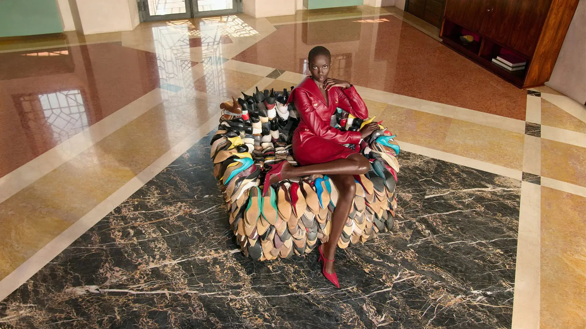 Model wearing a fitted red leather dress and matching pointed pumps while seated on a sculptural armchair made from stacked colorful shoes, photographed inside a marble-clad interior for the Ferragamo Fall/Winter 2026 campaign.