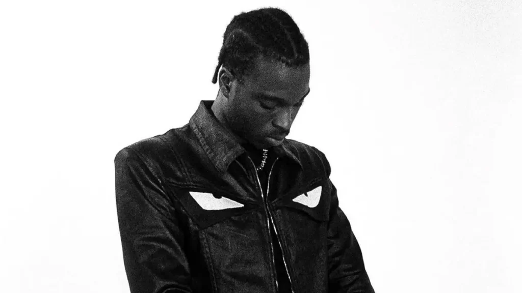 FimiGuerrero in a monochrome portrait, looking downward while wearing a dark jacket with bold eye-shaped graphic across the chest