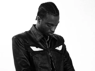 FimiGuerrero in a monochrome portrait, looking downward while wearing a dark jacket with bold eye-shaped graphic across the chest