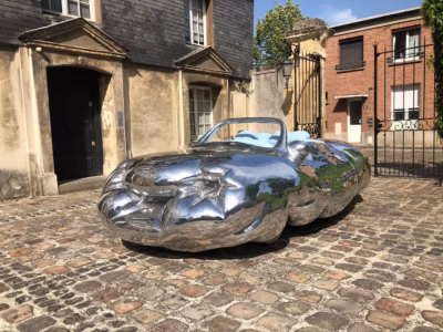 Mirror-polished sculpture convertible with light blue seats parked on a cobblestone courtyard outside historic stone buildings on a sunny day