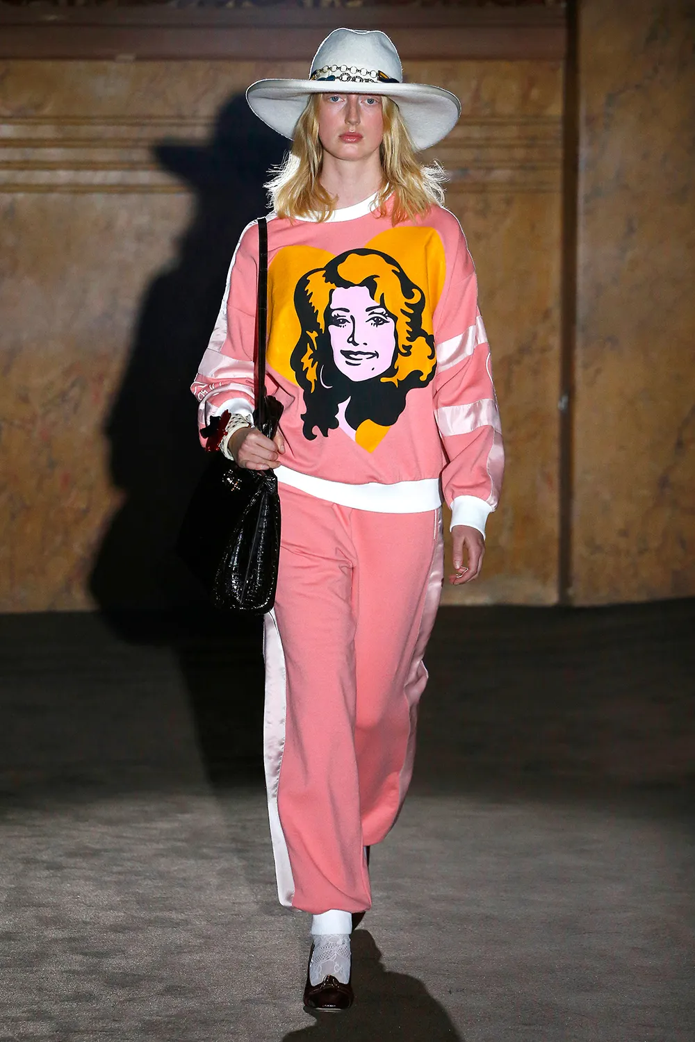 Model in a pink tracksuit with a Dolly Parton graphic, white cowboy hat, and black handbag on a runway