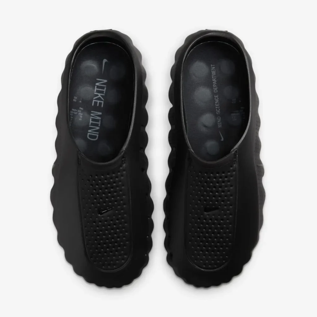 Top-down view of a pair of black Nike mules featuring perforated foot coverings, cushioned footbeds with Nike Mind branding, and chunky ripple-edge sole