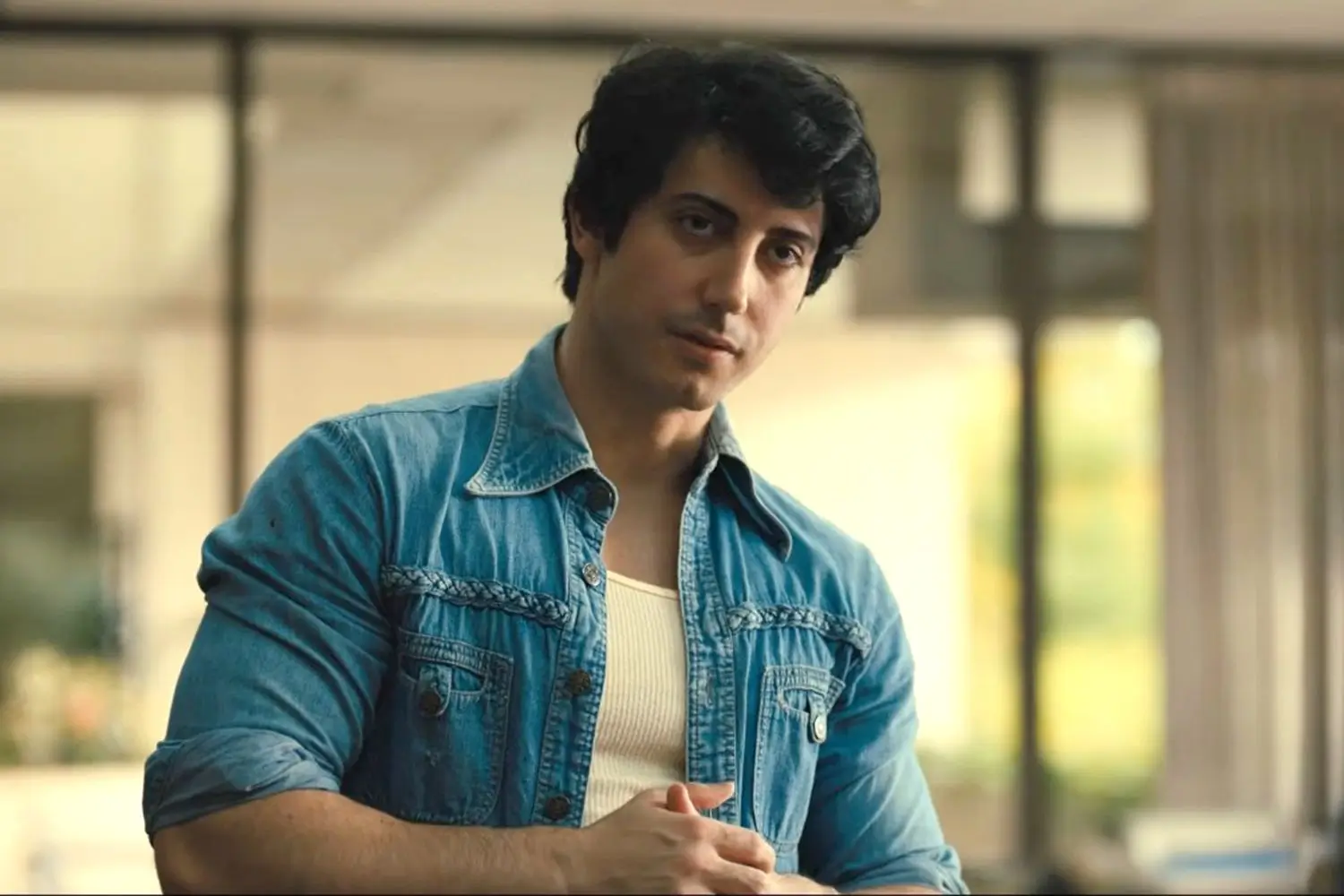 Man wearing a blue denim shirt over a white ribbed tank top in a softly lit interior.