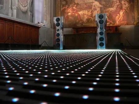 Pair of high-end floorstanding loudspeakers displayed on an illuminated LED floor inside a historic gallery, with Renaissance-style murals and ornate architectural details creating a dramatic contrast between classic art and contemporary audio design.