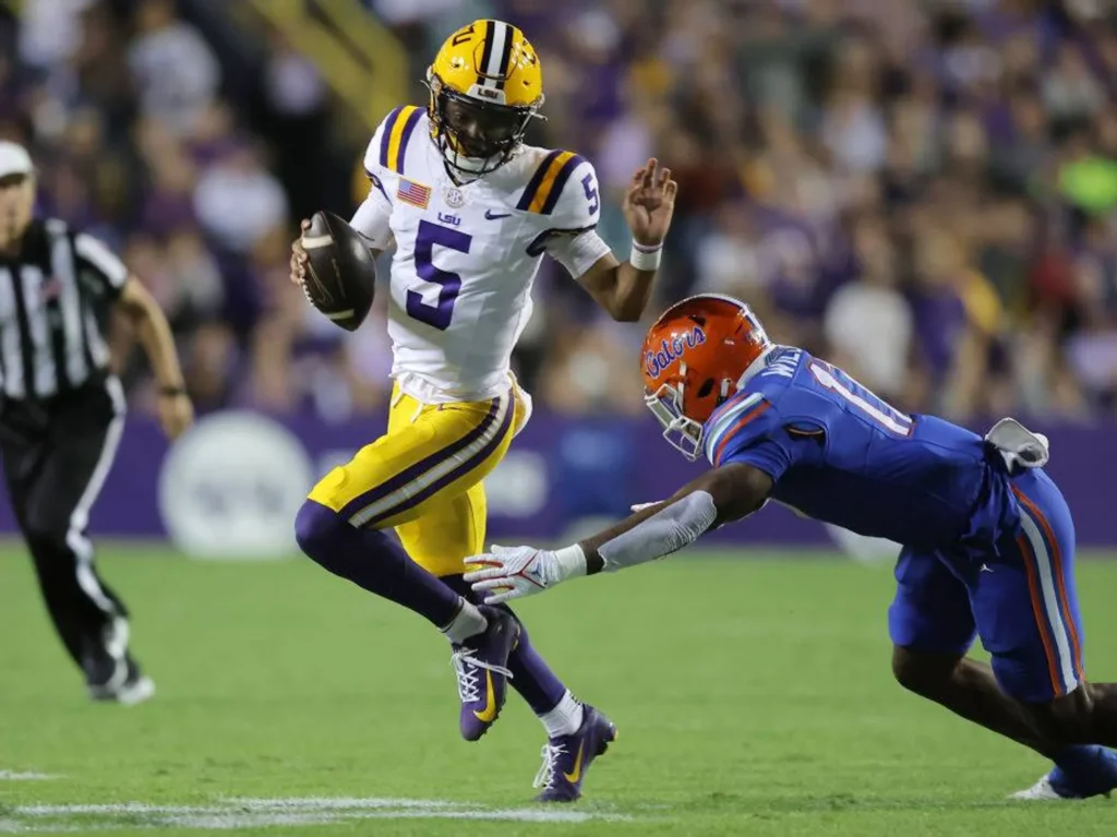 College football quarterback Jayden Daniels in a white LSU Tigers No. 5 jersey, yellow pants and yellow helmet runs with the football while evading a diving Florida Gators defender in a blue uniform and orange helmet during a night game