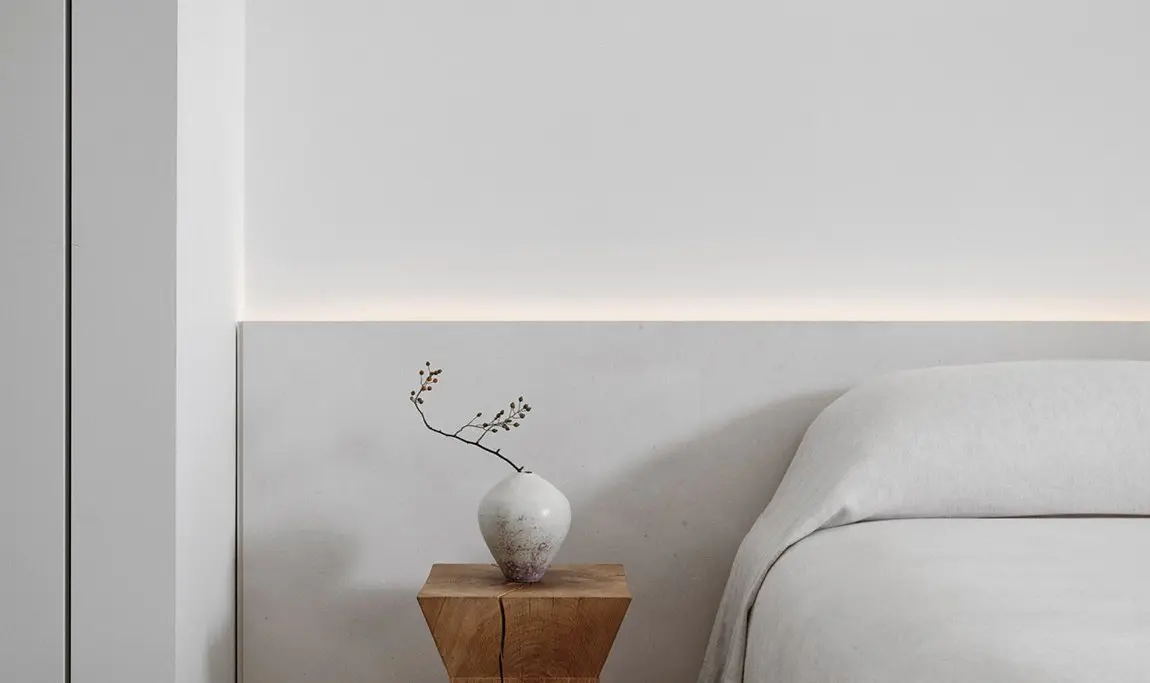 Minimalist bedroom with a white upholstered bed, warm concealed lighting, and a wooden bedside table topped with a ceramic vase and delicate branch.