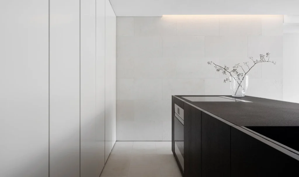Min kitchen featuring floor-to-ceiling white cabinet, texture white stone walls, a dark kitchen island, and a glass vase hold branch