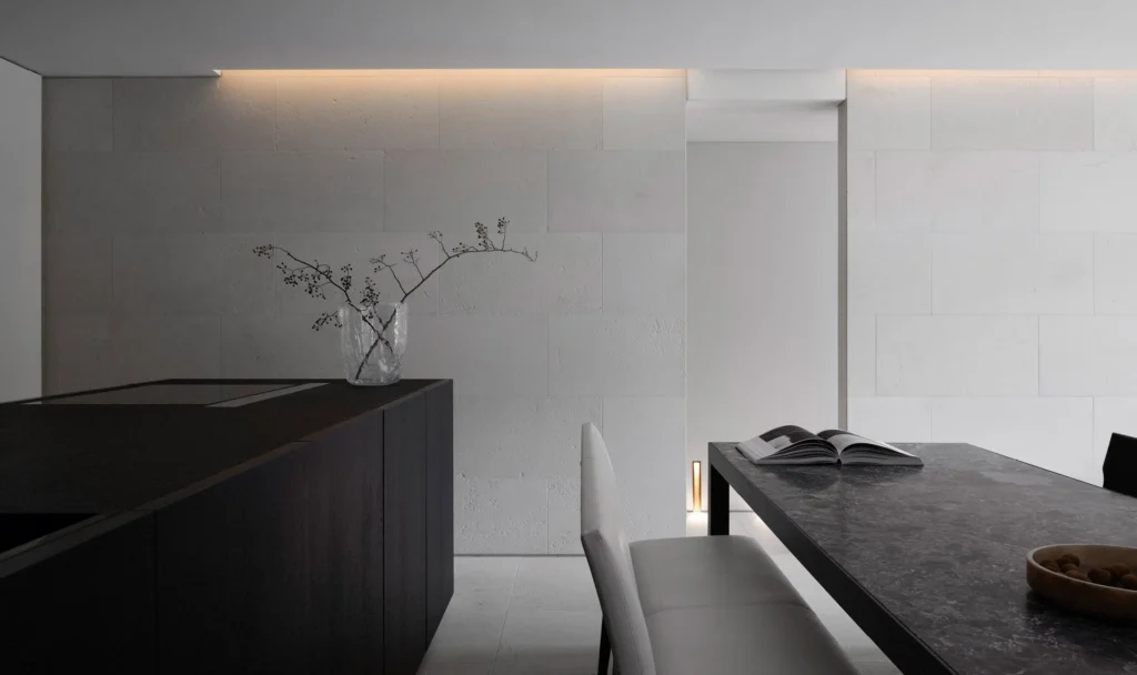 Wide view of a monochrome kitchen and dine space with a black island, dark stone table, white seat, texture walls, and subtle warm light