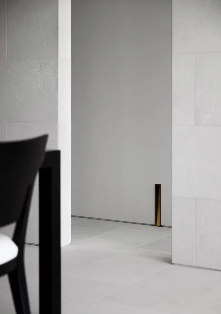 Architecture detail of a min white interior with textured stone walls, pale tiled flooring, dark furniture, and a narrow recess floor-level light