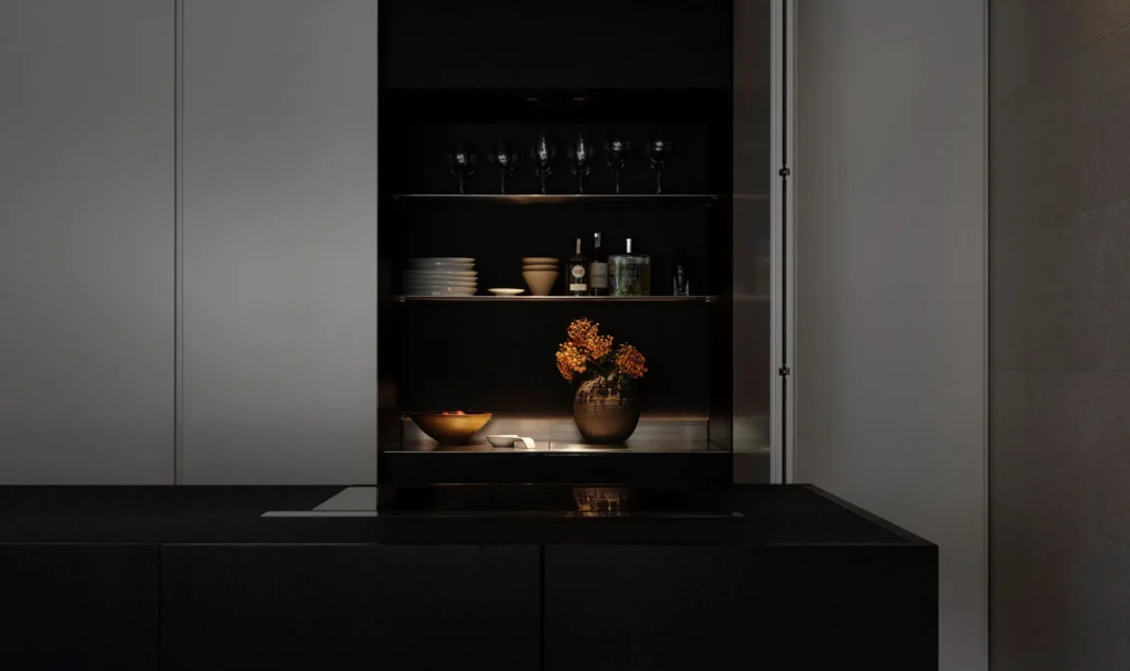 Moody min kitchen with a matte-black island and show open cabinet display glassware, ceramics, bottles, and a vase of orange flower