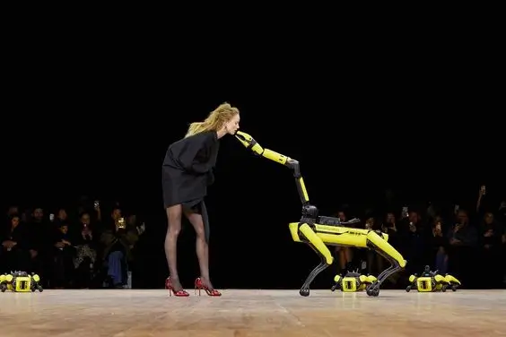A model in a black mini dress and red heels leans forward to touch noses with a yellow quadruped robot on a runway stage, while spectators watch from the audience.