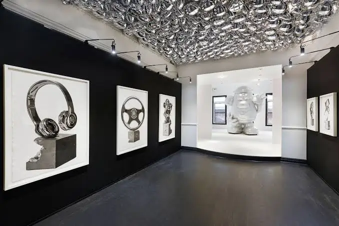 Contemporary Art Gallery With Black Walls, Framed Monochrome Drawings, A Reflective Sculptural Ceiling, And A Large White Character Sculpture.