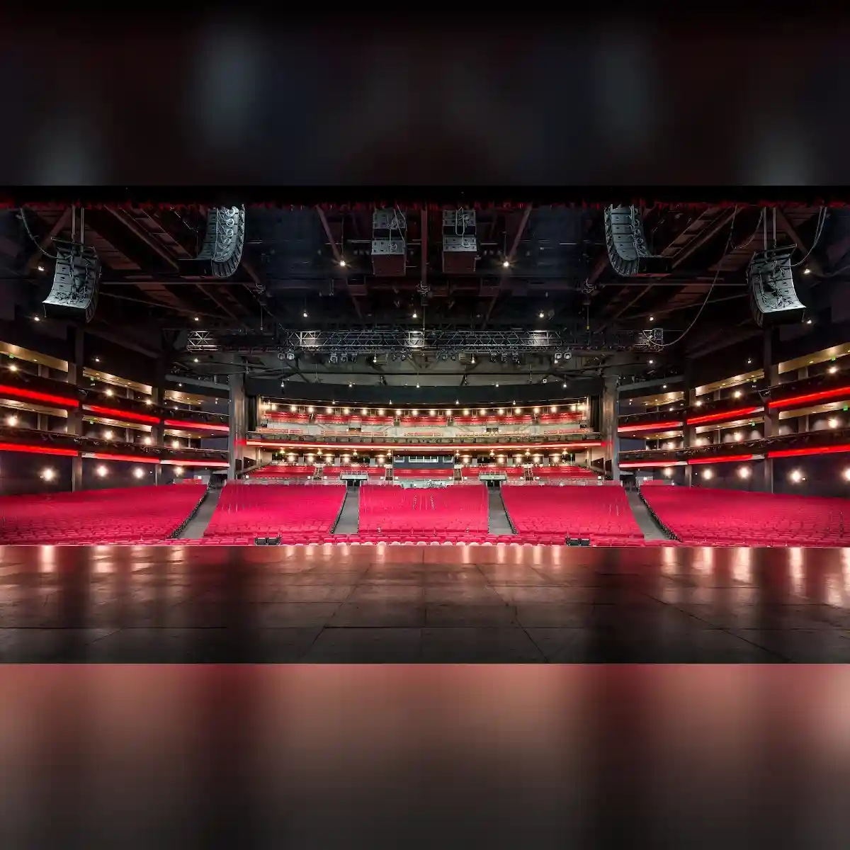 View From A Theater Stage Toward Rows Of Red Seats, Multiple Balconies, Overhead Speakers, And Stage Lighting