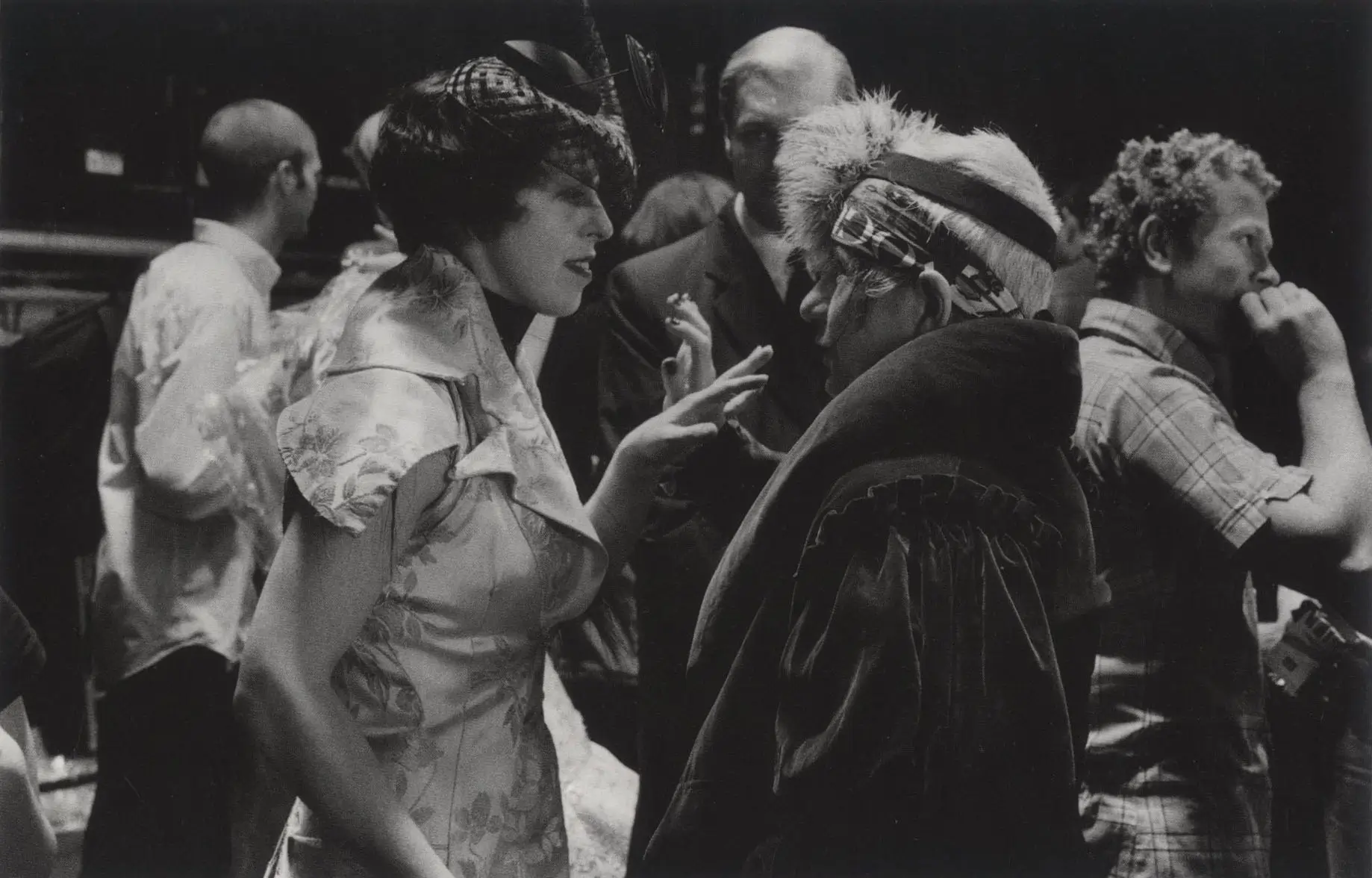 Two fashion figures in elaborate hats converse backstage as guests mingle in a black-and-white event photograph.