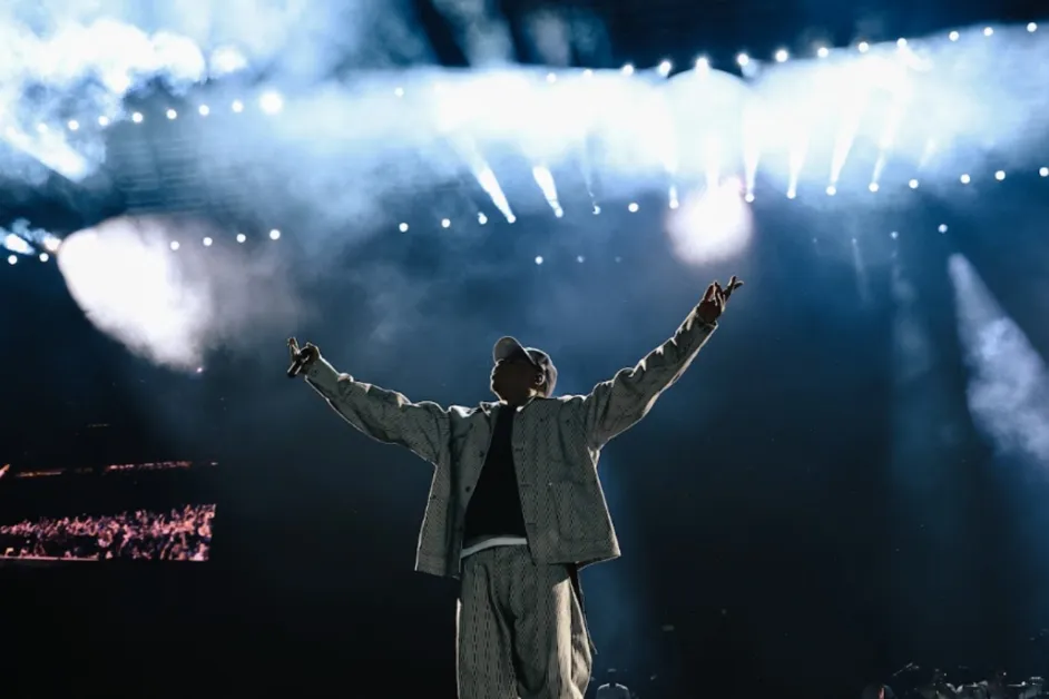 Jay-Z raises both arms beneath bright stage lights and smoke during a stadium performance