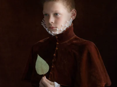 Young Person Wearing A Rich Brown Velvet Period-Style Garment With White Lattice Collar And Cuffs, Holding A Single Green Leaf Against A Dark Brown Background