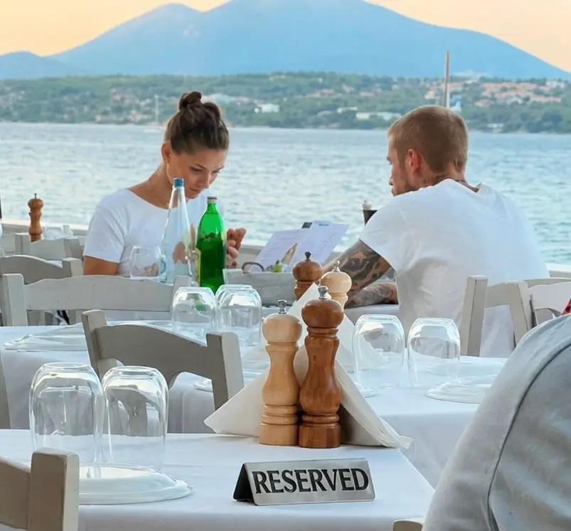 Hailey and Justin Bieber dining at a waterfront restaurant, seated across from each other with menus, surrounded by white tables overlooking the sea and distant mountains.