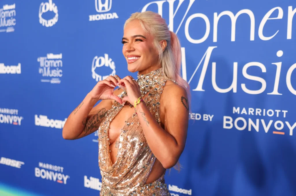 Smiling Karol Gin a sparkling gold halter gown forming a heart with her hands on the blue Billboard Women in Music red carpet