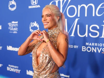 Smiling Karol Gin a sparkling gold halter gown forming a heart with her hands on the blue Billboard Women in Music red carpet