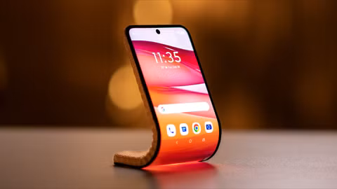 Concept smartphone with a flexible OLED display bending into a self-supporting curved shape on a tabletop, illuminated by warm ambient lighting.