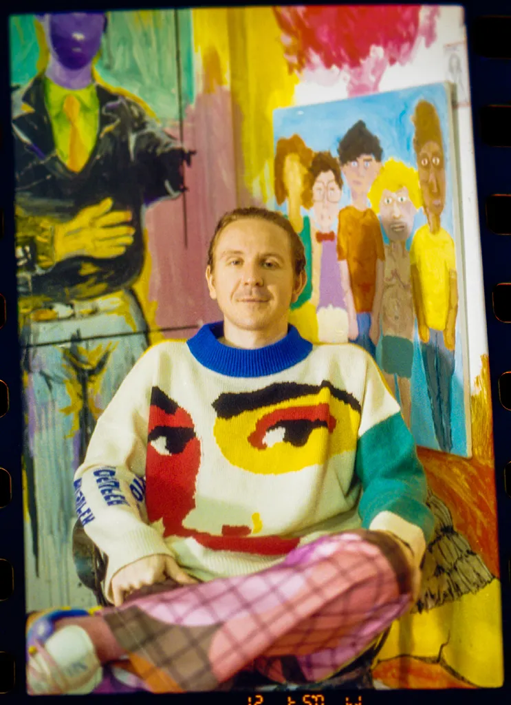 Artist Seated In Front Of Colorful Figurative Paintings Wearing A Graphic Knit Sweater And Pink Plaid Trousers