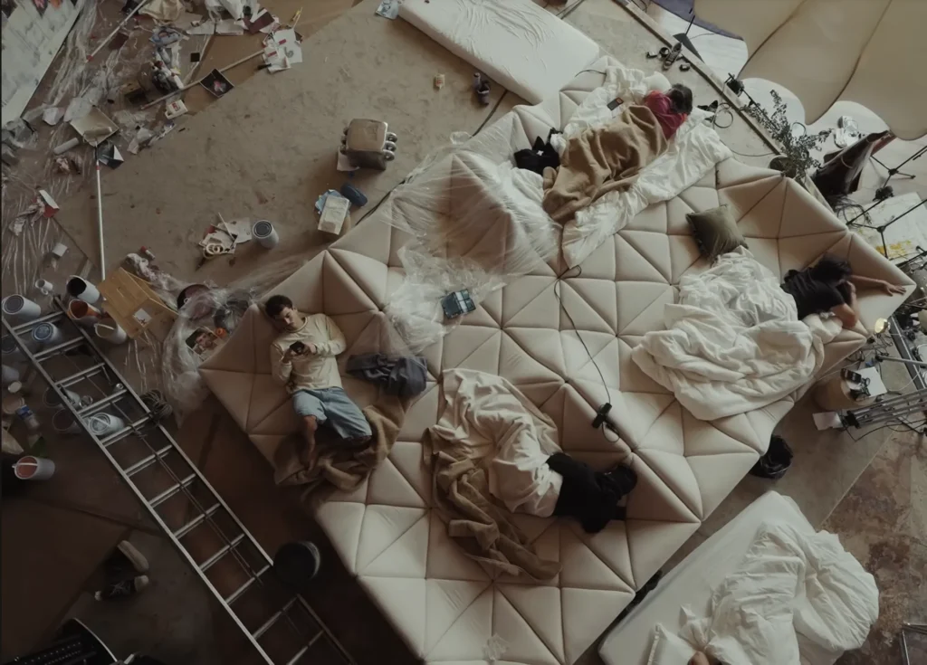 Overhead view of a sprawl cream geometric sofa covered with blankets, electronics, and people relaxing inside a clutter creative studio