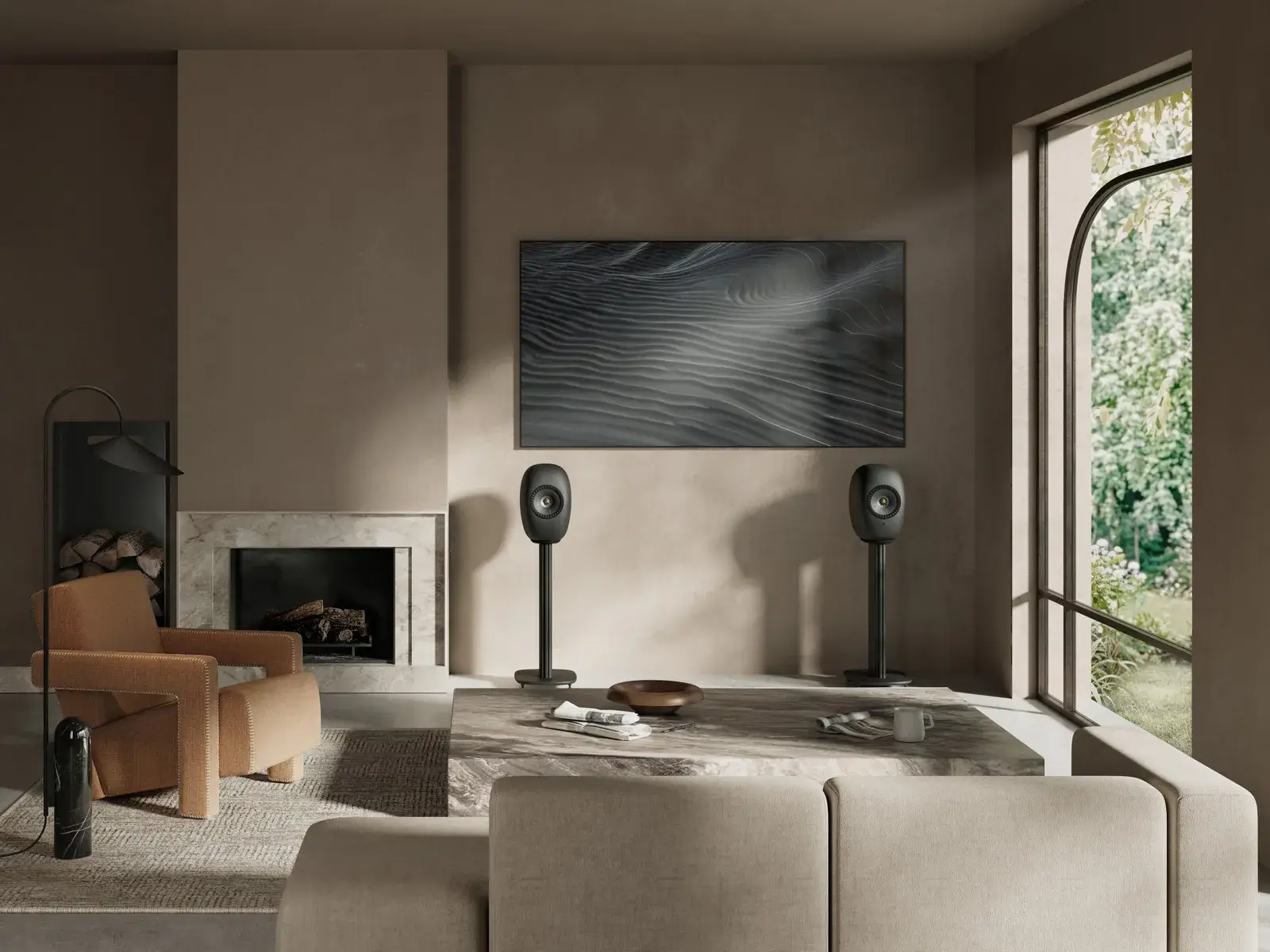 Modern living room featuring a pair of matte black KEF LSX II LT wireless speakers on matching floor stands, positioned beneath a wall-mounted television beside a fireplace and framed by minimalist furnishings with abundant natural light.