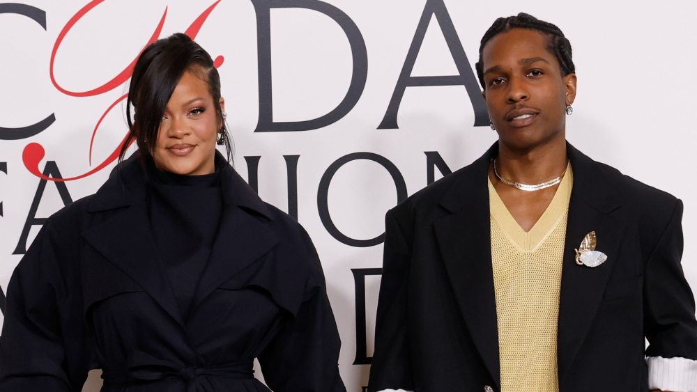 Two fashion figures pose together on a red carpet in coordinated black tailoring, accented with refined jewelry and layered styling against an event backdrop.