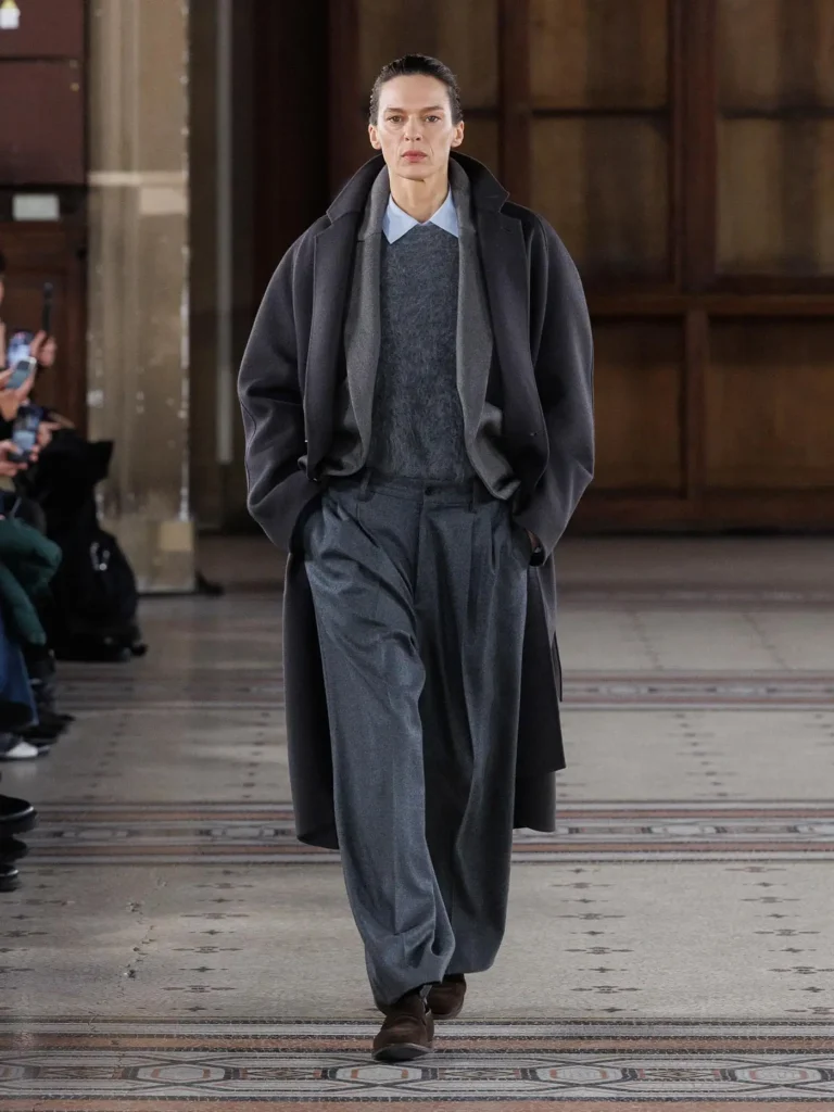 Model wearing a charcoal oversized overcoat layered over a grey knit sweater and pale blue collared shirt, paired with wide-leg grey trousers and brown loafers during an indoor runway show