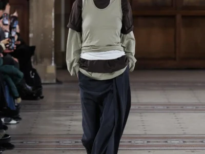 Model wearing layered olive, black, and white tops with relaxed navy trousers, brown suede loafers, and slouchy styling in a minimalist runway look