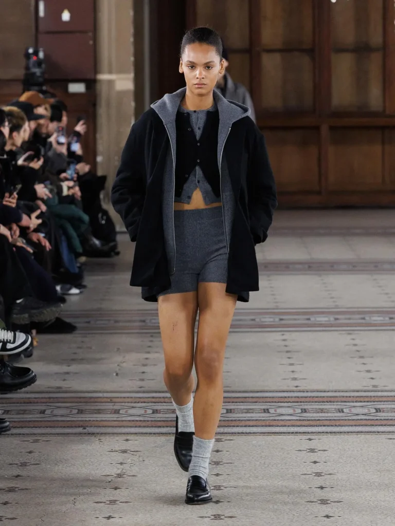 Model wearing a black oversized jacket over layered grey knit separates and fitted shorts, finished with grey socks and black loafers on the runway