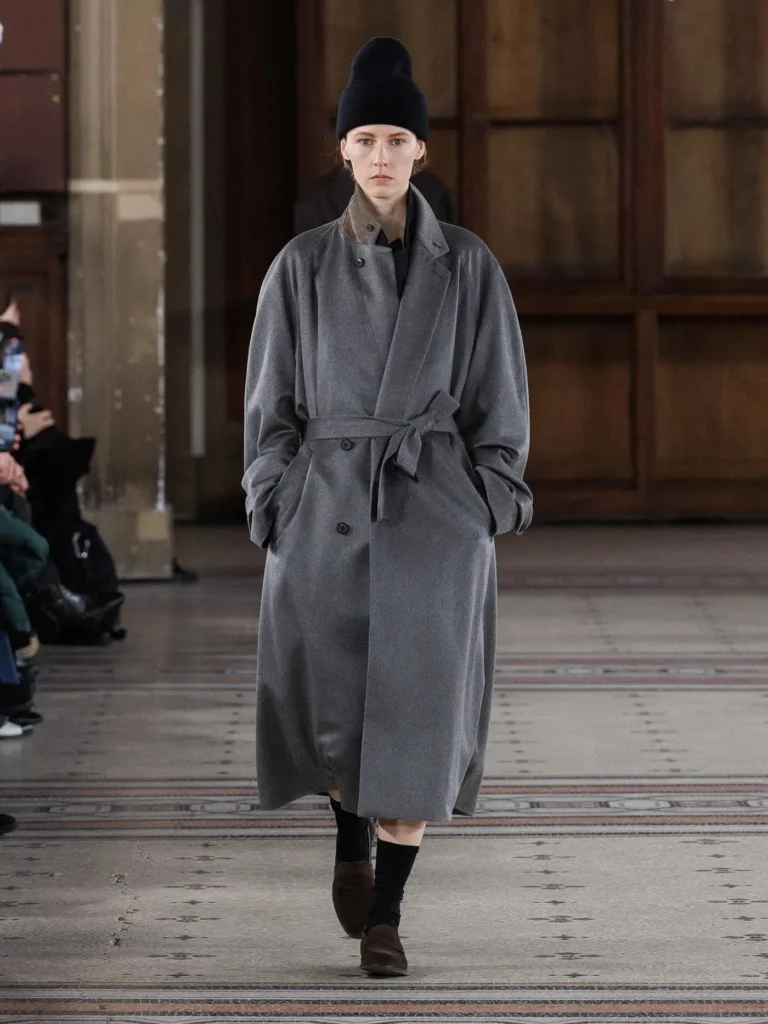 Model wearing a belted grey wool overcoat with a black knit beanie, black socks, and brown loafers in a clean, understated runway ensemble