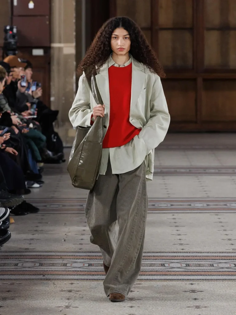 Model wearing an oversized light grey blazer over a vibrant red sweater and wide taupe trousers, carrying a large olive leather shoulder bag