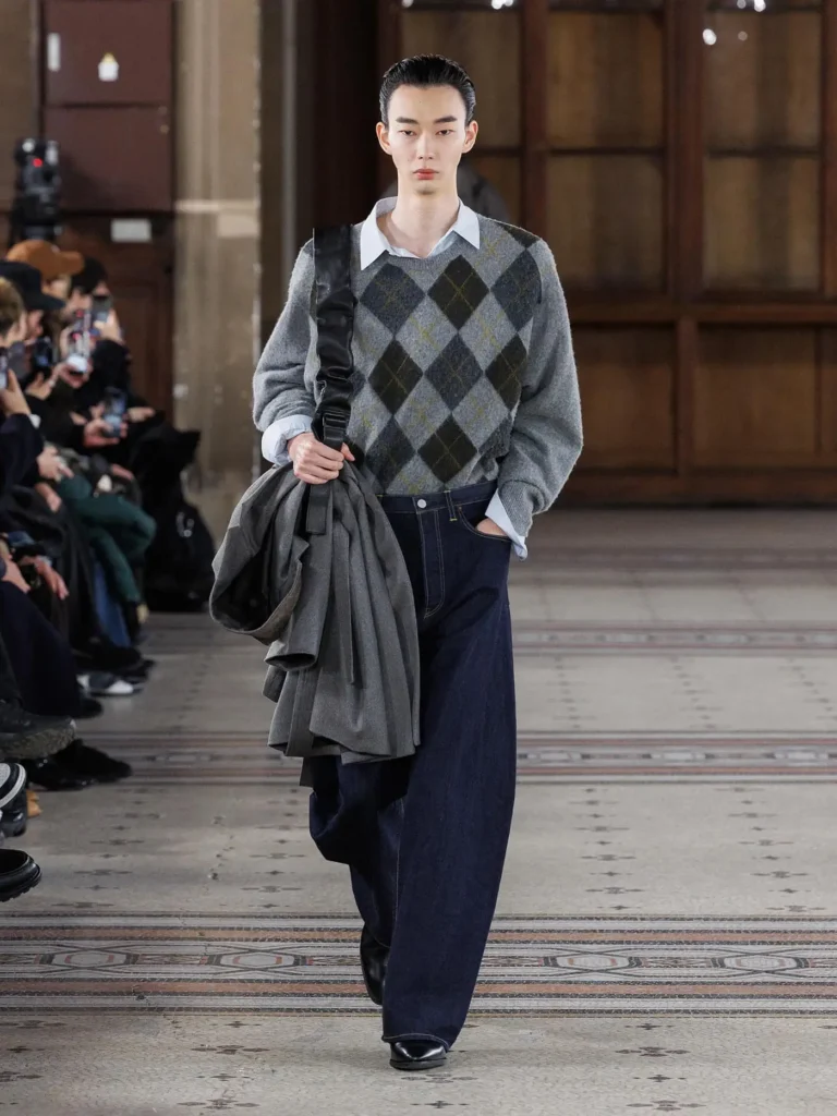 Model wearing a grey argyle sweater over a pale blue collared shirt with wide dark denim trousers, carrying a draped grey coat over one shoulder