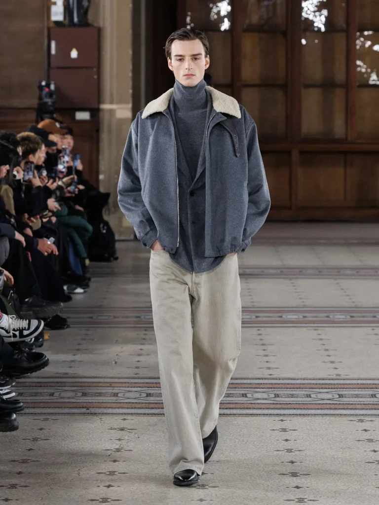 Model wearing a grey jacket with a shearling collar over layered knitwear, paired with cream wide-leg trousers and black leather shoe