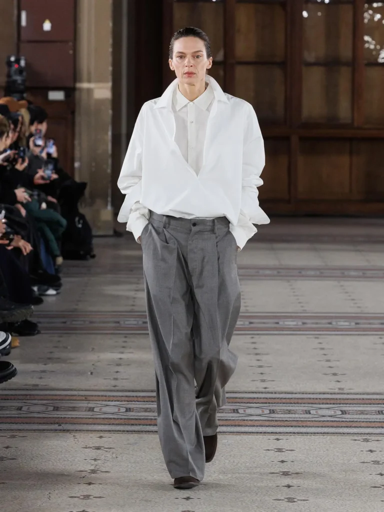 Model wearing an oversized white wrap-front shirt tucked into pleated grey trousers with brown suede shoes in a refined runway look