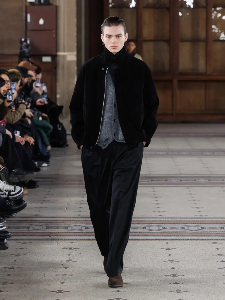 Model wearing a black zip-front jacket layered over grey knit separates with relaxed black trousers and brown suede shoe