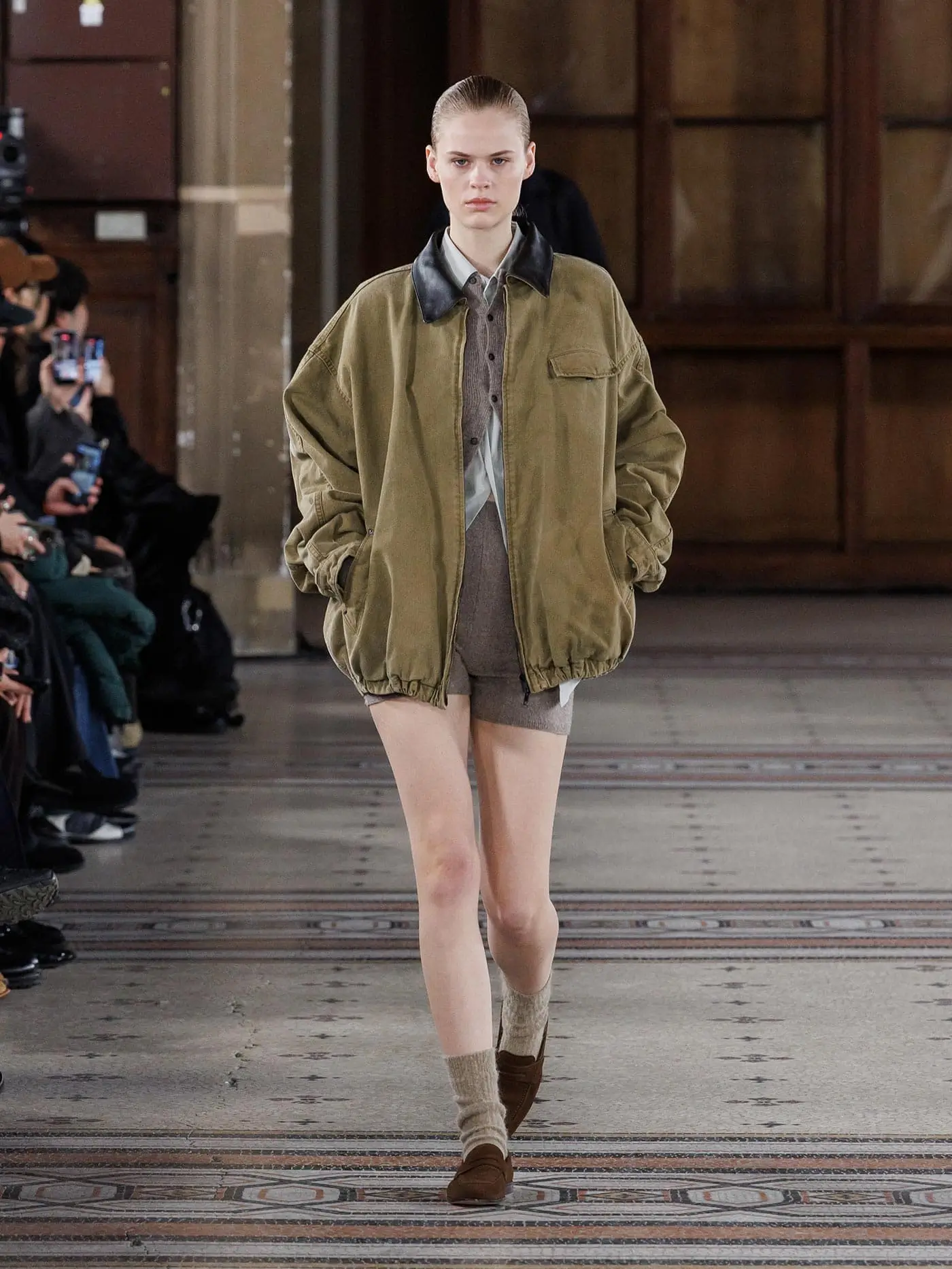 Model walking the runway in an oversized olive bomber jacket with a black leather collar, layered knit separates, ankle socks, and brown loafers.