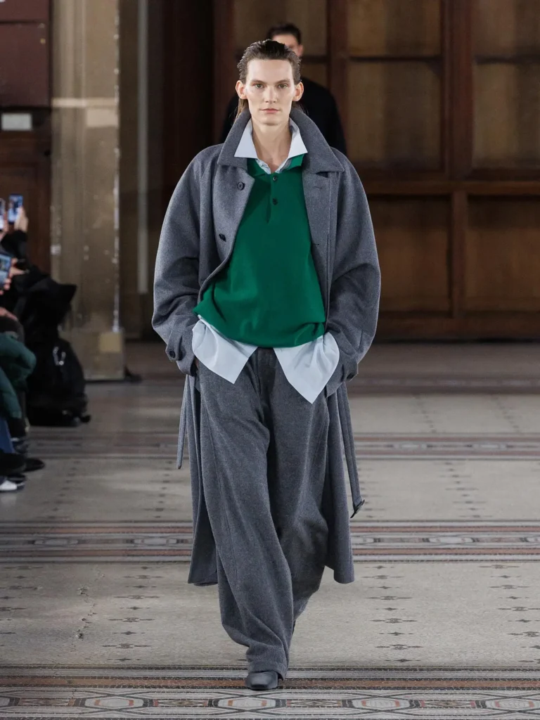 Model wearing a long grey overcoat layered over a green knit rugby-style sweater, oversized white shirt, and relaxed grey trousers in a contemporary tailored runway look
