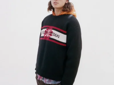 Person wear a black Stüssy Design knit sweater, striped green beanie, sunglasses, orange hood, and brown short