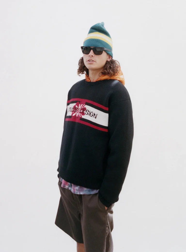 Person wear a black Stüssy Design knit sweater, striped green beanie, sunglasses, orange hood, and brown short