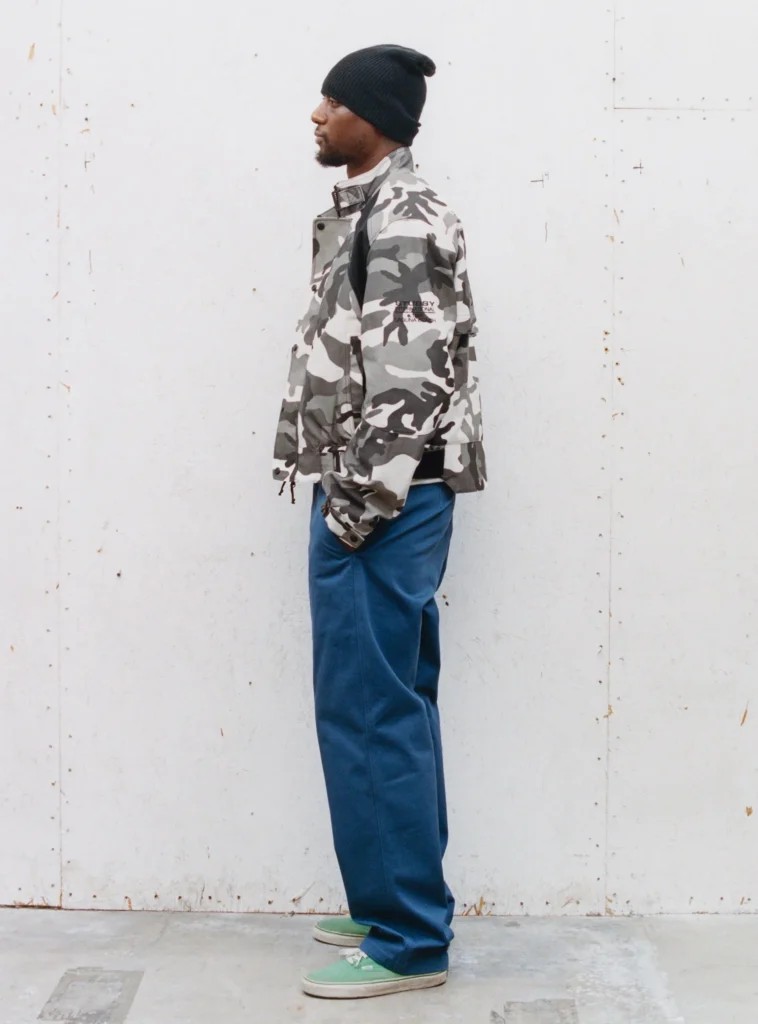 Person stands in profile wearing a black beanie, gray camouflage jacket, wide blue pants, and green shoe
