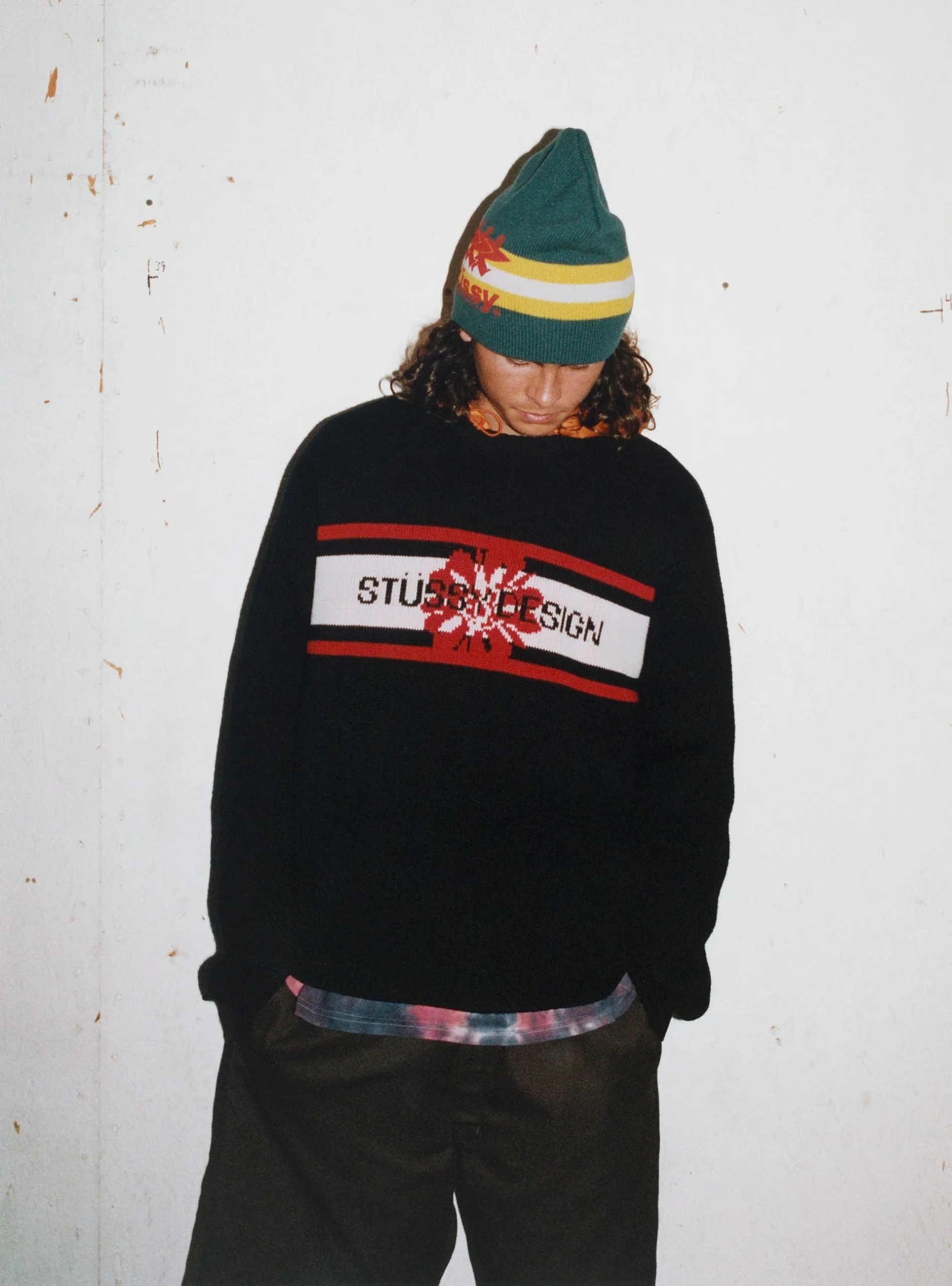 Person Looks Down While Wearing A Black Stüssy Design Knit Sweater And Green Striped Beanie