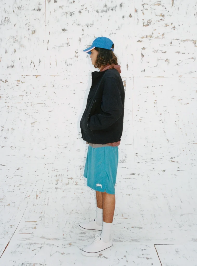 Person stands in profile wear a blue cap, dark jacket, turquoise shorts, white sock, and white shoe