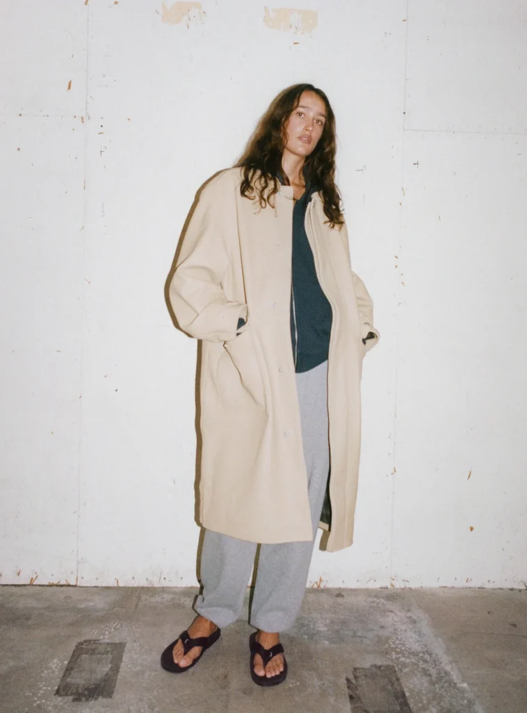 Person wear a long cream coat over a dark top and gray pant with black sandal