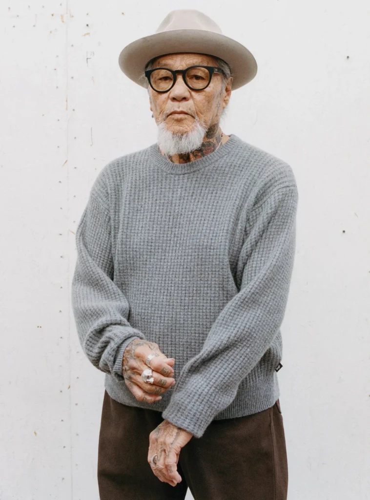 Older person wear a gray texture sweater, brown pants, wide-brim hat, black glasses, and silver ring